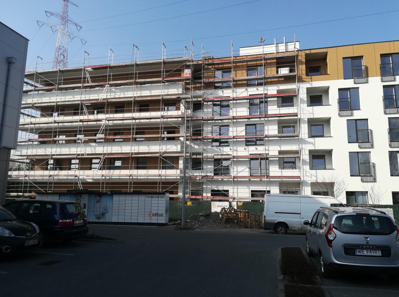 Wielopiętrowy budynek w trakcie prac elewacyjnych, widoczne rusztowania na części ściany, fragmenty elewacji w kolorze białym i brązowym, na pierwszym planie Paczkomat InPost i zaparkowane samochody.