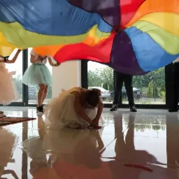 Dzieci w sukienkach bawią się pod wielobarwną chustą animacyjną w jasnym pomieszczeniu z dużymi oknami.