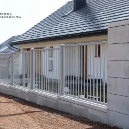 Szare ogrodzenie palisadowe z pionowym profilem 2x2 cm i prześwitem 11 cm, zamontowane na bloczkach Roma Mega w kolorze snow, na tle domu z ciemnym dachem.