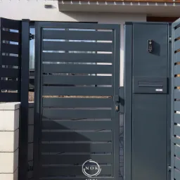 Grafitowa furtka palisadowa z subtelnymi prześwitami, wbudowanym domofonem i skrzynką na listy, tworząca nowoczesne wejście do domu. Widoczny fragment ogrodzenia.