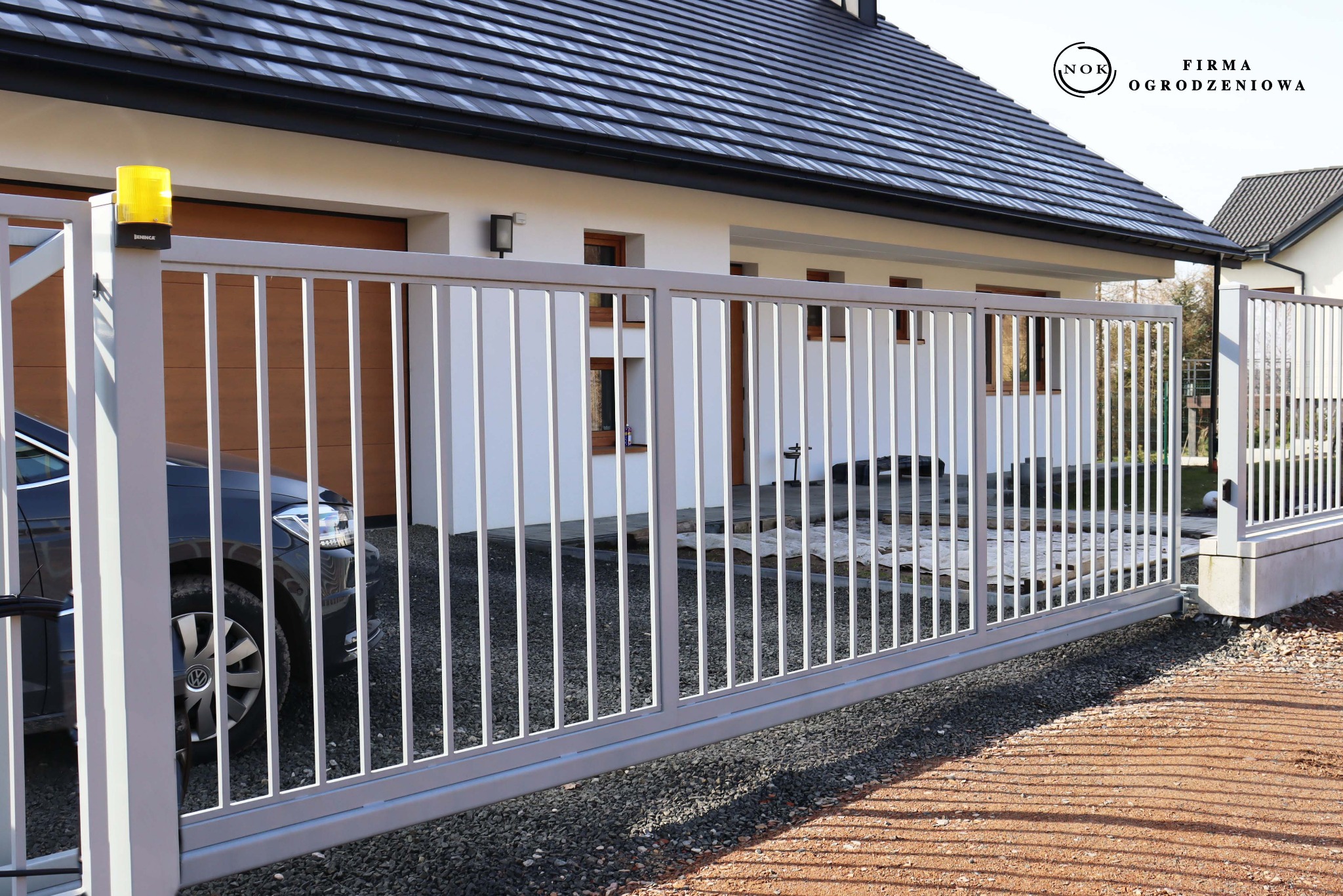 Szara brama przesuwna palisadowa (profil 2x2 cm, prześwit 11 cm) zamontowana do bloczków Roma Mega, snow. Widoczny fragment samochodu i sygnalizator.