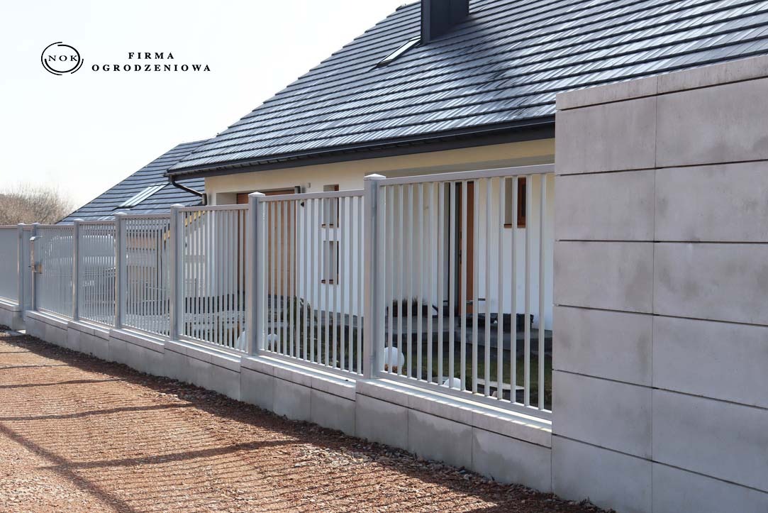 Szare ogrodzenie palisadowe z pionowym profilem 2x2 cm i prześwitem 11 cm, zamontowane na bloczkach Roma Mega w kolorze snow, na tle domu z ciemnym dachem.