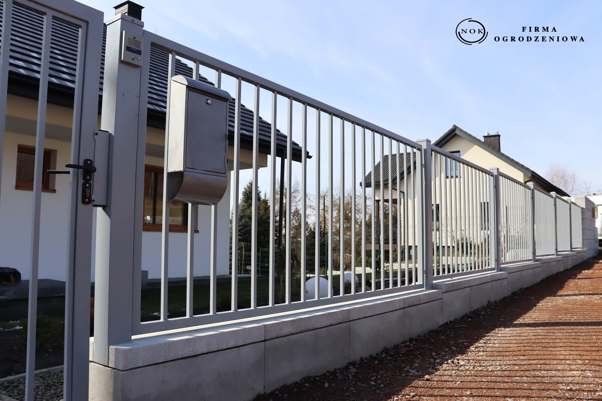 Szare ogrodzenie palisadowe z profilem 2x2 cm, prześwit 11 cm, w pionie, kolor RAL7040. Montowane na bloczkach Roma Mega, snow. Widok z perspektywy.