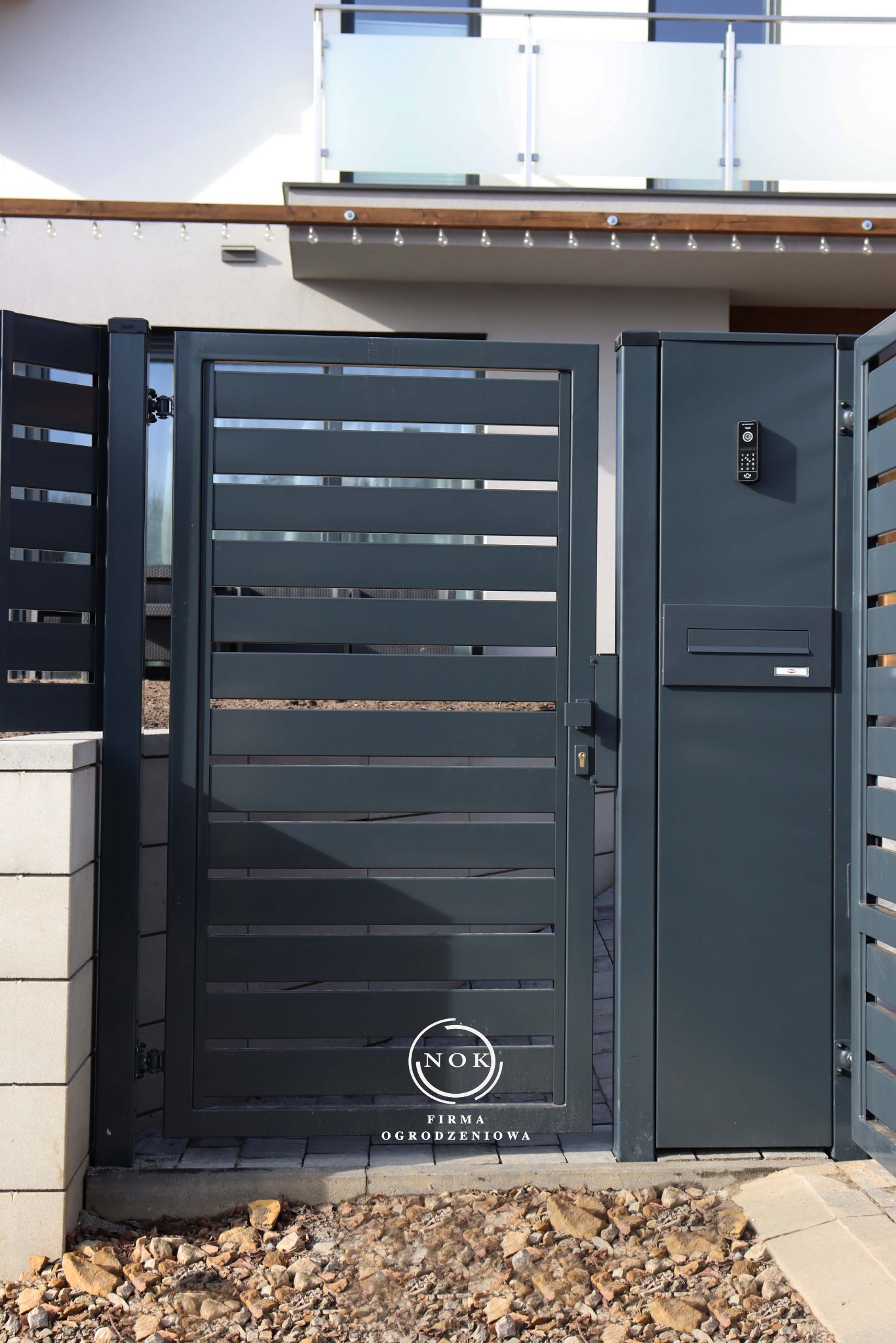 Grafitowa furtka palisadowa z subtelnymi prześwitami, wbudowanym domofonem i skrzynką na listy, tworząca nowoczesne wejście do domu. Widoczny fragment ogrodzenia.