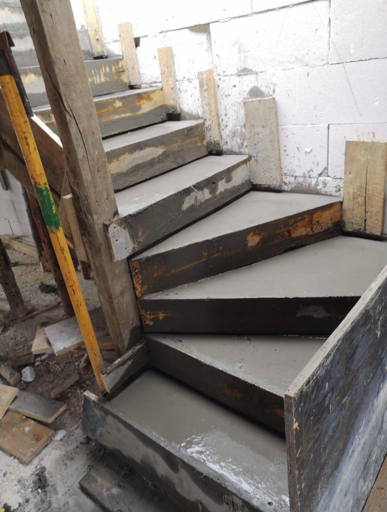 Surowe betonowe schody w drewnianym szalunku, poziomica przymocowana do boku konstrukcji, ściana z bloczków betonowych w tle.
