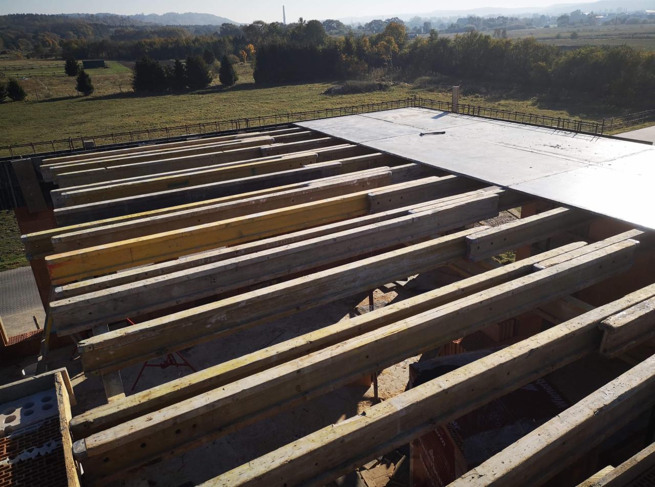 Wiązary dachowe z drewnianych belek i szalunku na płaskim dachu w trakcie budowy, widok z góry na tle zielonego krajobrazu.