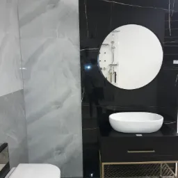 Nowoczesna łazienka z umywalką nablatową, okrągłym lustrem i marmurowymi płytkami w odcieniach szarości i czerni, akcentowana złotymi elementami w szafce pod umywalką.