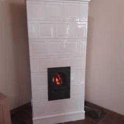 Biały piec kaflowy z czarnymi drzwiczkami, płomienie widoczne wewnątrz, drewno ułożone obok, na podłodze z ciemnych płytek.