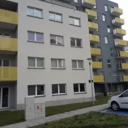 Nowoczesny budynek wielorodzinny z szarą i białą elewacją, żółtymi balkonami, widok z poziomu chodnika, zaparkowany biały samochód na niebieskim miejscu parkingowym.