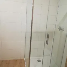 Szklana kabina prysznicowa z chromowanymi profilami i odpływem liniowym, białe płytki na ścianach, podłoga wyłożona panelami imitującymi drewno.