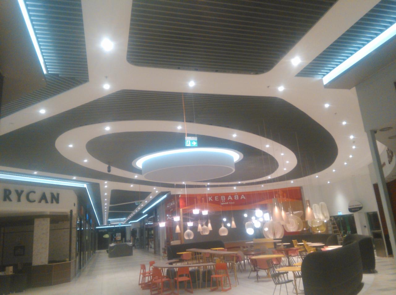Sufit rastrowy w centrum handlowym z okrągłymi elementami dekoracyjnymi i oświetleniem LED, widok na strefę restauracyjną z meblami.