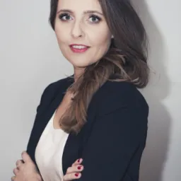 Portret kobiety z ciemnymi włosami i czerwonymi ustami, ubrana w czarny żakiet i białą bluzkę, pozująca z założonymi rękami na tle jasnej ściany.