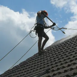 Pracownik w kasku i uprzęży, myje ciśnieniowo dach pokryty ciemną dachówką, na tle błękitnego nieba z białymi chmurami.