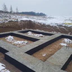 Zalane betonem ławy fundamentowe w zimowej scenerii, widoczne wystające pręty zbrojeniowe i śnieg w wykopie.