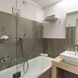 Nowoczesna łazienka z wanną oddzieloną szklanym panelem, szare płytki, biała ceramika i minimalistyczna armatura, w tle półka z ręcznikami.