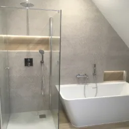 Nowoczesna łazienka na poddaszu z kabiną prysznicową ze szklanymi drzwiami, wolnostojącą białą wanną, szarą płytką imitującą jodełkę na ścianach oraz wnęką z oświetleniem LED.