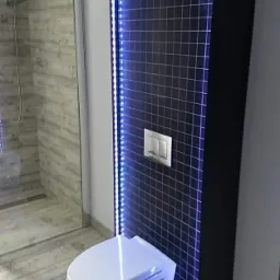 Nowoczesna toaleta podwieszana z czarną ścianką obłożoną drobną mozaiką, oświetlona niebieskimi diodami LED, widoczna w łazience z prysznicem.