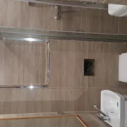 Wykończona łazienka z kabiną prysznicową ze szklanymi drzwiami, umywalką z szafką i podwieszaną toaletą na tle płytek imitujących drewno; widoczne też pralka i akcesoria montażowe.