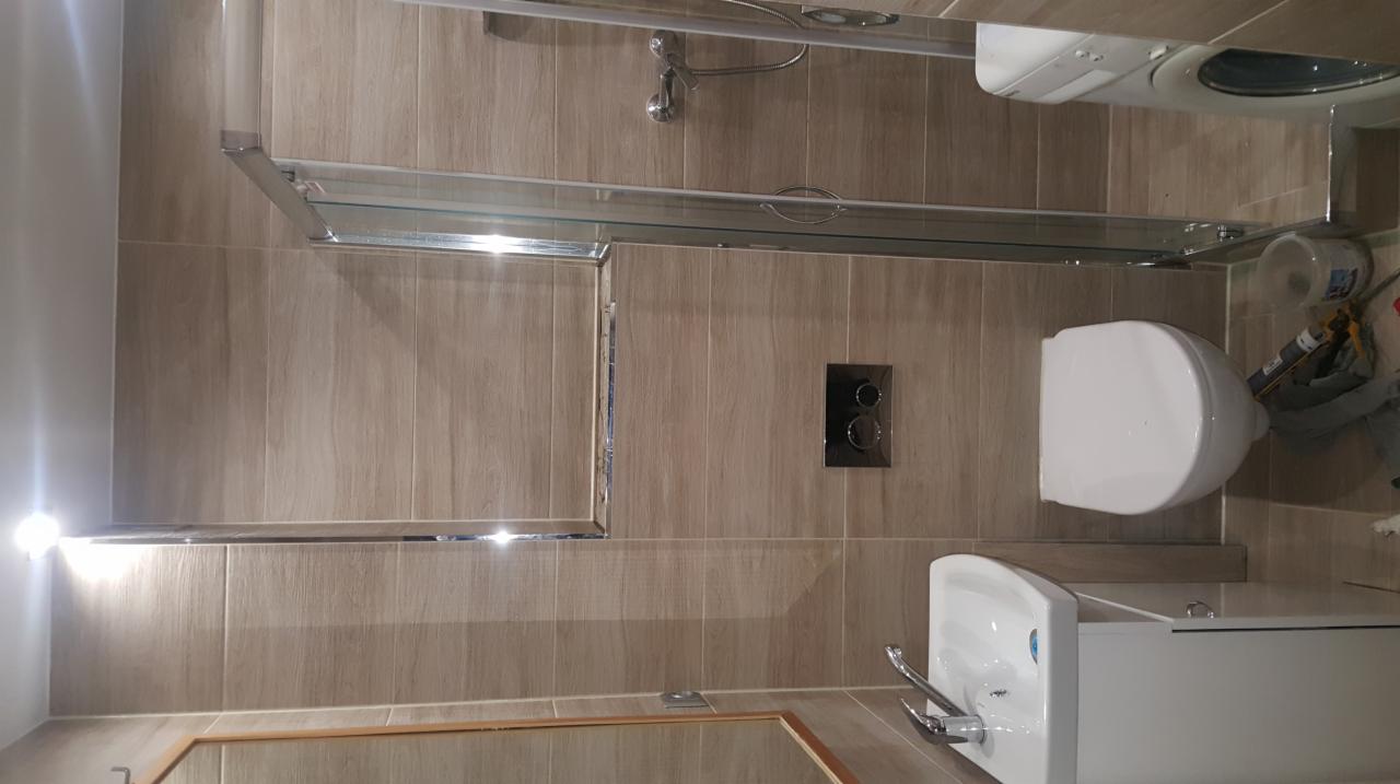 Wykończona łazienka z kabiną prysznicową ze szklanymi drzwiami, umywalką z szafką i podwieszaną toaletą na tle płytek imitujących drewno; widoczne też pralka i akcesoria montażowe.