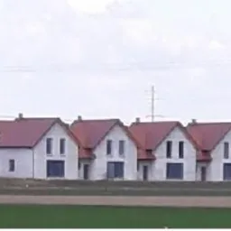 Szereg identycznych domów jednorodzinnych w różnym stadium budowy, od surowego stanu do wykończonych elewacji, z czerwonymi dachami i białymi ścianami, widok z daleka na tle pochmurnego nieba.