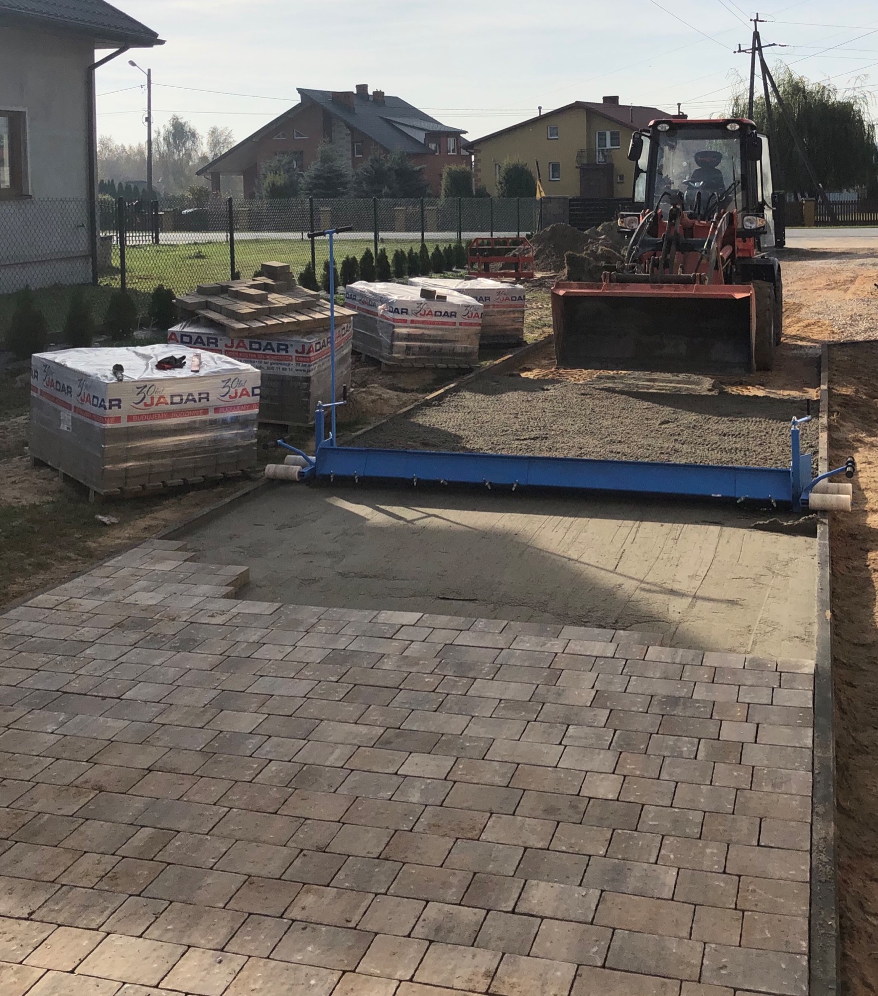 Częściowo ułożona, brązowo-szara kostka brukowa na posesji, palety z kostką Jadar, ubijarka do piasku i traktor z łyżką w tle.