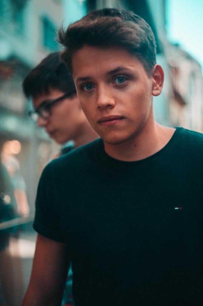 Portret młodego mężczyzny o niebieskich oczach w czarnym T-shircie z logo Tommy Hilfiger, drugi mężczyzna z okularami w tle.