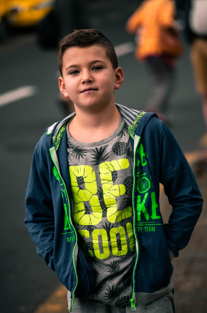 Portret chłopca w bluzie z kapturem i koszulce z nadrukiem, stojącego na ulicy z rękami w kieszeniach.