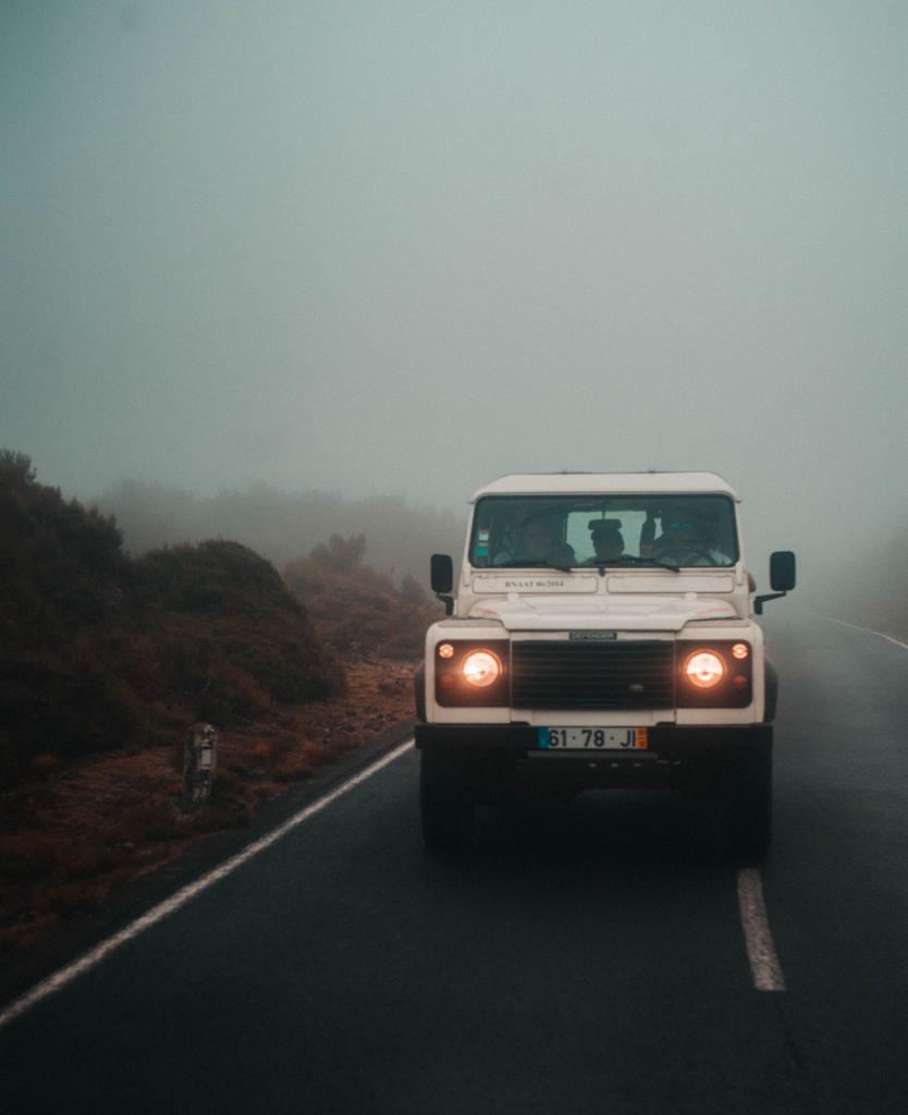 Biały Land Rover Defender z włączonymi światłami mijania jedzie po mokrej, asfaltowej drodze we mgle, widok z przodu.
