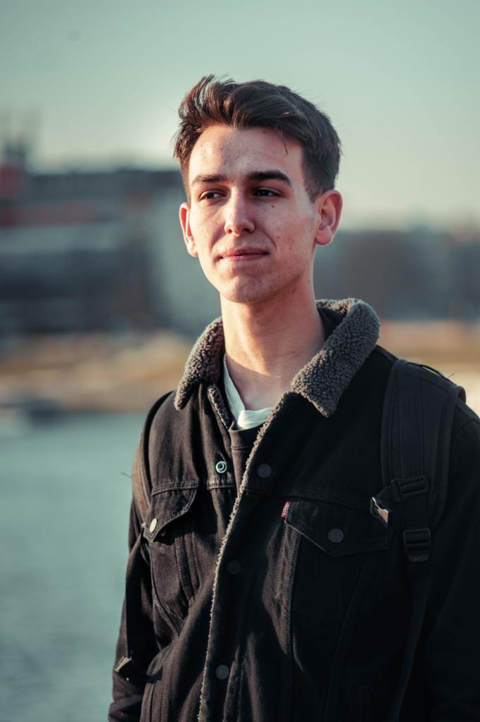 Portret młodego mężczyzny w kurtce z kożuszkiem, z plecakiem, spoglądającego w dal na tle miejskiego krajobrazu i rzeki.