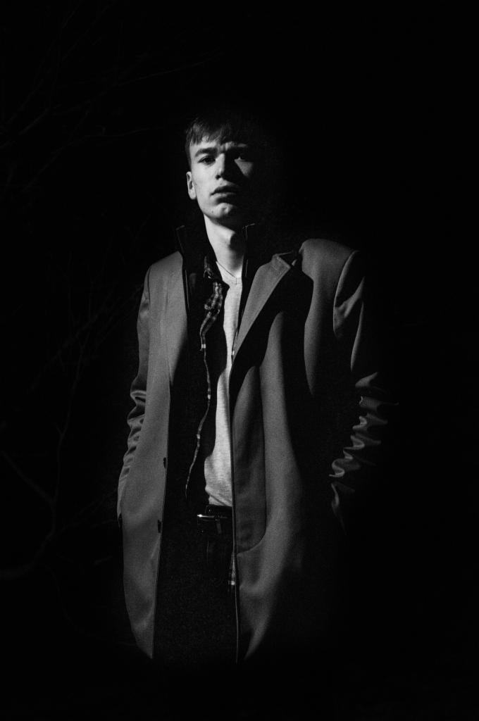 Portret młodego mężczyzny w płaszczu, z ciemnym tłem i mocnym oświetleniem podkreślającym rysy twarzy.