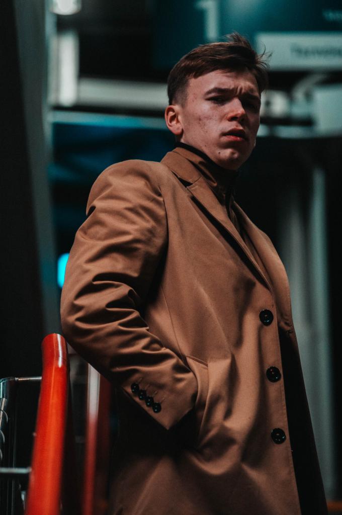 Młody mężczyzna w brązowym płaszczu i czarnym golfie, pozujący na tle miejskiej architektury z czerwonymi poręczami i neonowym oświetleniem.