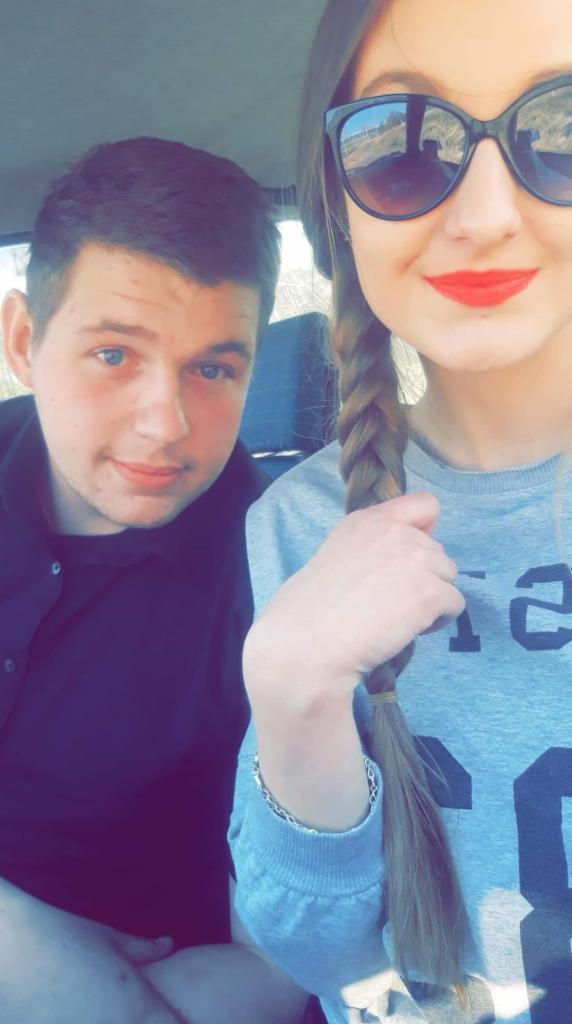 Autoportret pary w samochodzie: mężczyzna z niebieskimi oczami w czarnej koszuli, kobieta w okularach przeciwsłonecznych i czerwonej szmince, trzymająca w dłoni warkocz.