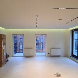 Usługi budowlane Zatorski Krzysztof - Wnętrze nowego mieszkania z wykończonymi ścianami i podłogą, widoczne kartony, grzejniki, balkony i nowoczesne oświetlenie sufitowe.