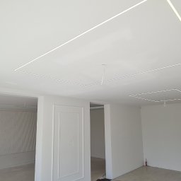 Usługi budowlane Zatorski Krzysztof - Wnętrze w trakcie wykańczania: białe ściany, sufit z podświetlanymi kwadratowymi wnękami, dekoracyjne panele ścienne, widoczne przewody i niedokończone detale.