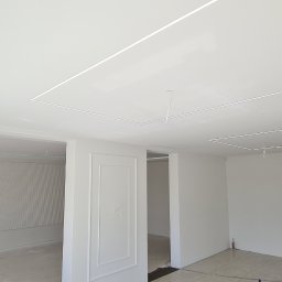 Usługi budowlane Zatorski Krzysztof - Wykończone wnętrze z białymi ścianami, ozdobnymi panelami ściennymi i podwieszanym sufitem z wbudowanym oświetleniem liniowym.