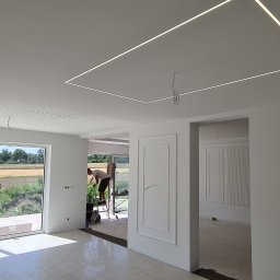 Usługi budowlane Zatorski Krzysztof - Wnętrze w trakcie wykańczania: białe ściany, wbudowane oświetlenie LED w suficie, pracownik na drabinie przy oknie, widok na zielone pola za oknem.