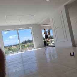 Usługi budowlane Zatorski Krzysztof - Jasne wnętrze w trakcie wykańczania, widoczny pracownik na drabinie, świeżo pomalowane ściany i sufit z nowoczesnym oświetleniem liniowym.