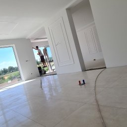 Usługi budowlane Zatorski Krzysztof - Jasne wnętrze w trakcie wykańczania, widoczny pracownik na drabinie malujący ścianę, ozdobne sztukaterie na ścianach, podłoga z połyskiem, nowoczesne oświetlenie sufitowe.