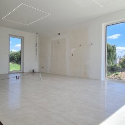 Usługi budowlane Zatorski Krzysztof - Wnętrze w trakcie wykańczania: beżowe płytki podłogowe, białe ściany z częściowo nałożoną gładzią i zaznaczonymi miejscami pod instalacje, kwadratowe panele LED w suficie.