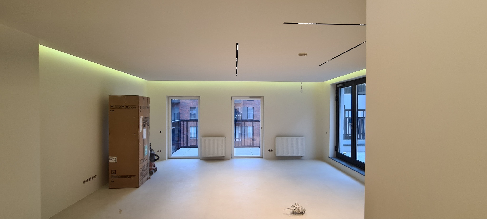 Wnętrze nowego mieszkania z wykończonymi ścianami i podłogą, widoczne kartony, grzejniki, balkony i nowoczesne oświetlenie sufitowe.