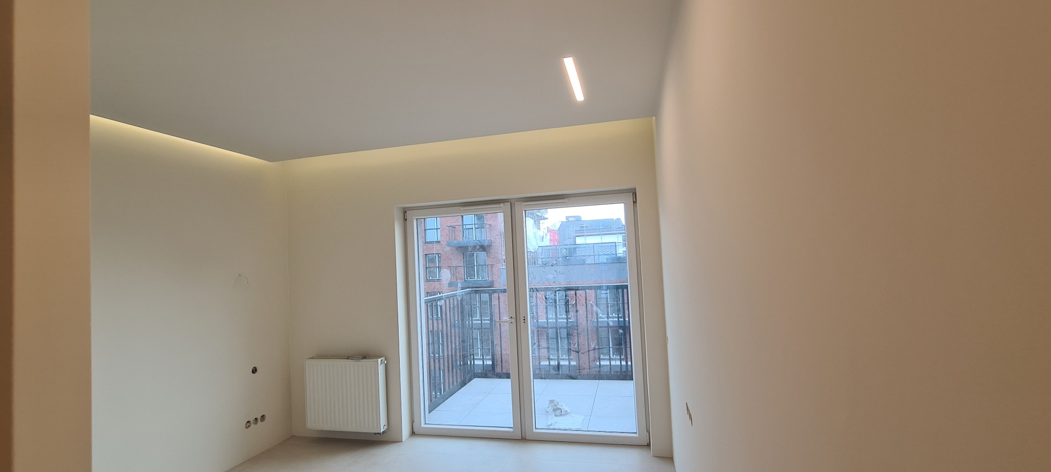 Jasne wnętrze po remoncie z widokiem na balkon, z minimalistycznym oświetleniem LED pod sufitem i na ścianie, widoczne otwory po gniazdkach elektrycznych.