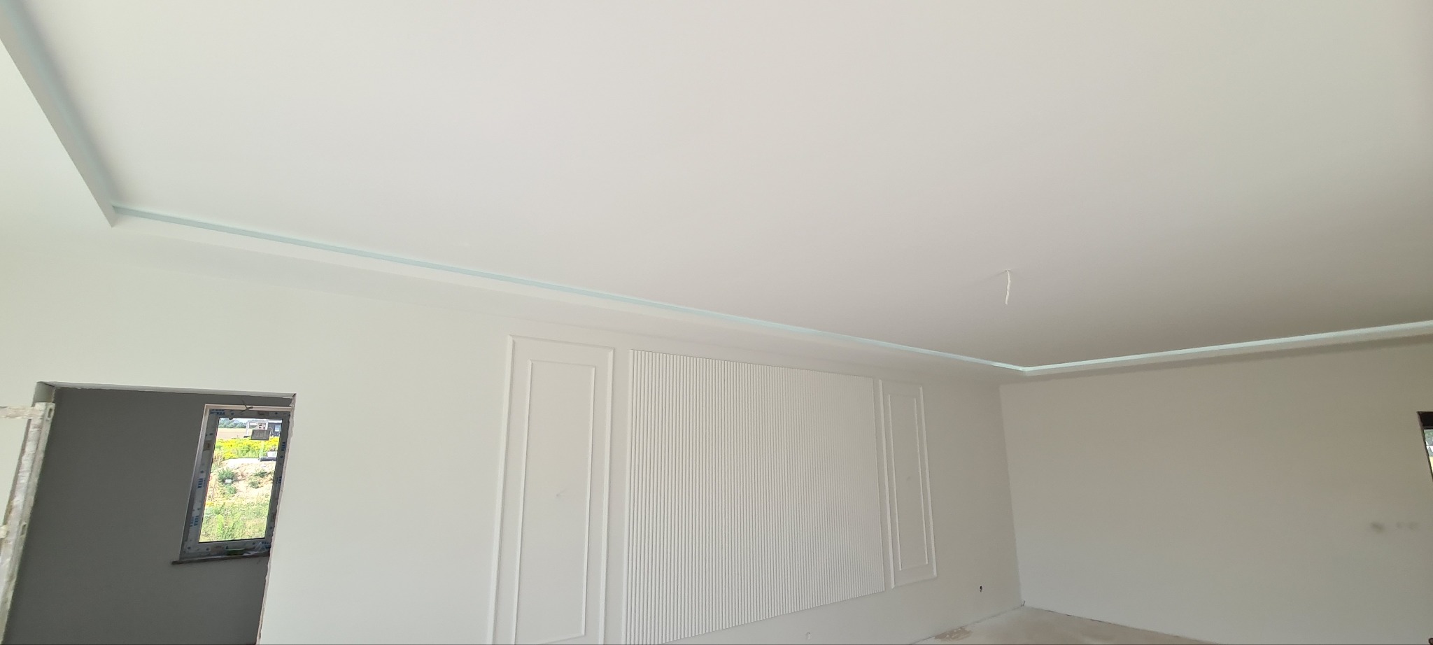 Wnętrze po remoncie z białą ścianą z dekoracyjnymi panelami i listwami oświetleniowymi LED przy suficie, widoczny otwór okienny z widokiem na zewnątrz.