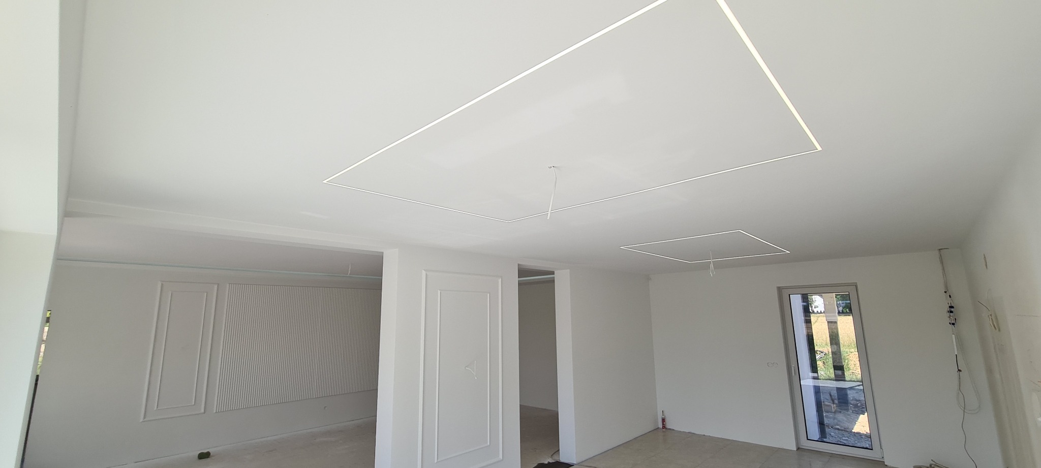 Wnętrze w trakcie wykańczania: białe ściany, sufit z podświetlanymi kwadratowymi wnękami, dekoracyjne panele ścienne, widoczne przewody i niedokończone detale.