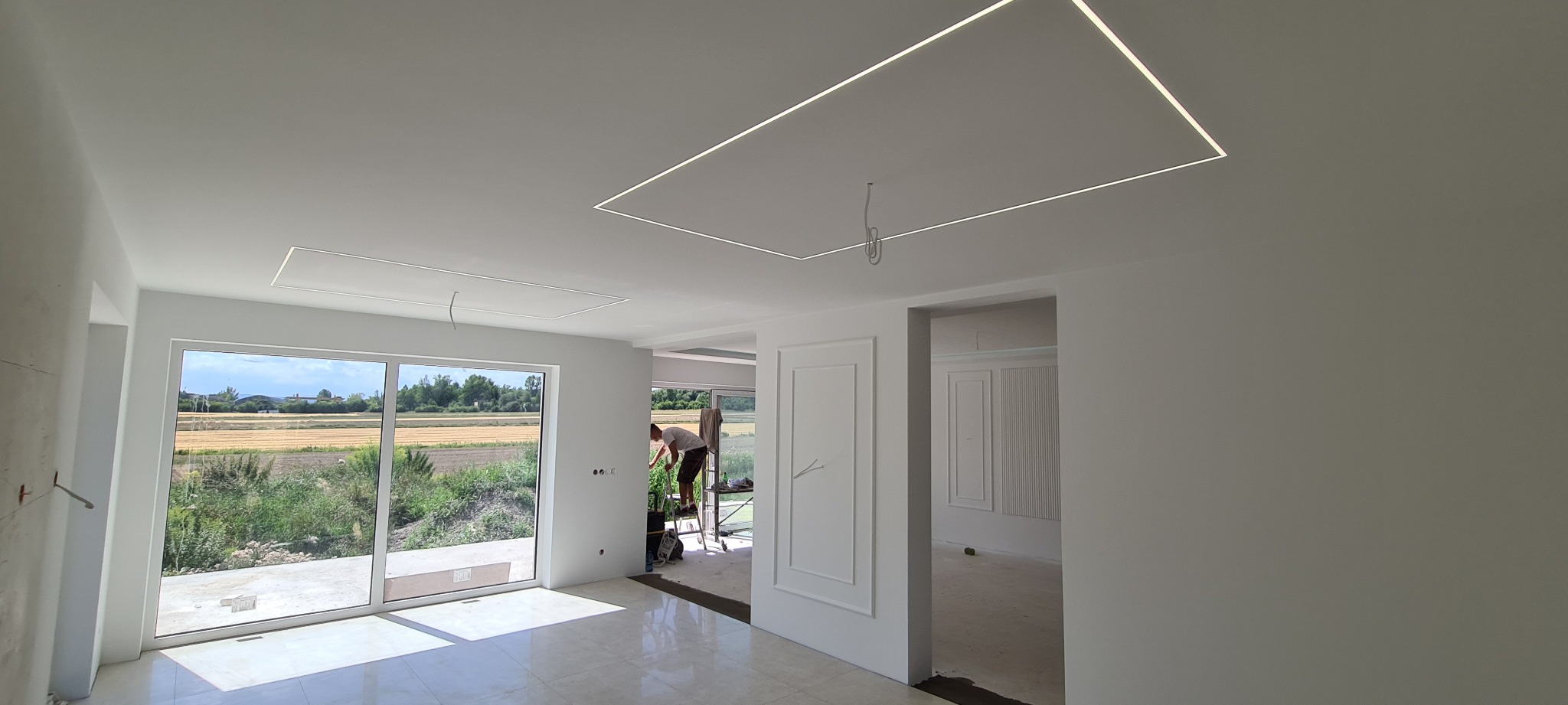 Wnętrze w trakcie wykańczania: białe ściany, wbudowane oświetlenie LED w suficie, pracownik na drabinie przy oknie, widok na zielone pola za oknem.