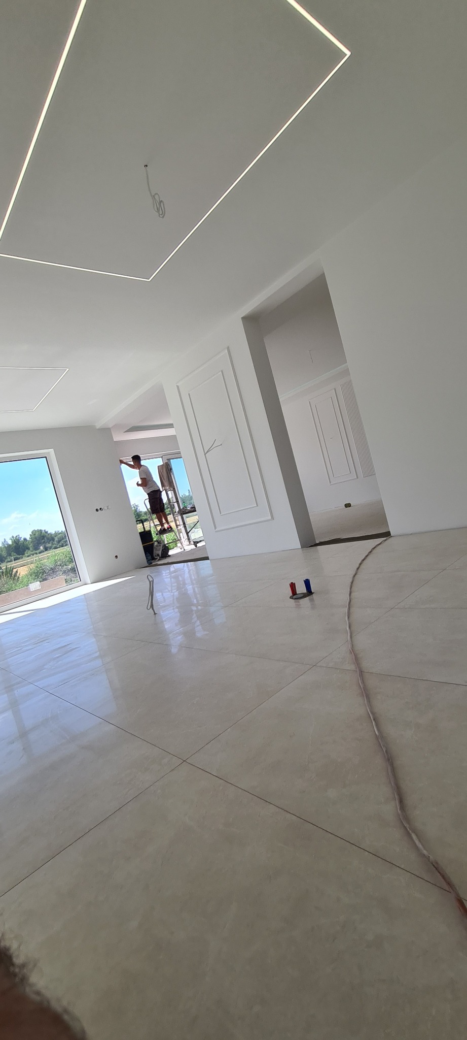 Jasne wnętrze w trakcie wykańczania, widoczny pracownik na drabinie malujący ścianę, ozdobne sztukaterie na ścianach, podłoga z połyskiem, nowoczesne oświetlenie sufitowe.