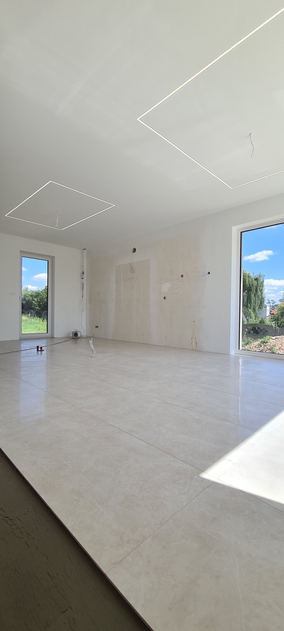 Wnętrze w trakcie wykańczania: beżowe płytki podłogowe, białe ściany z częściowo nałożoną gładzią i zaznaczonymi miejscami pod instalacje, kwadratowe panele LED w suficie.