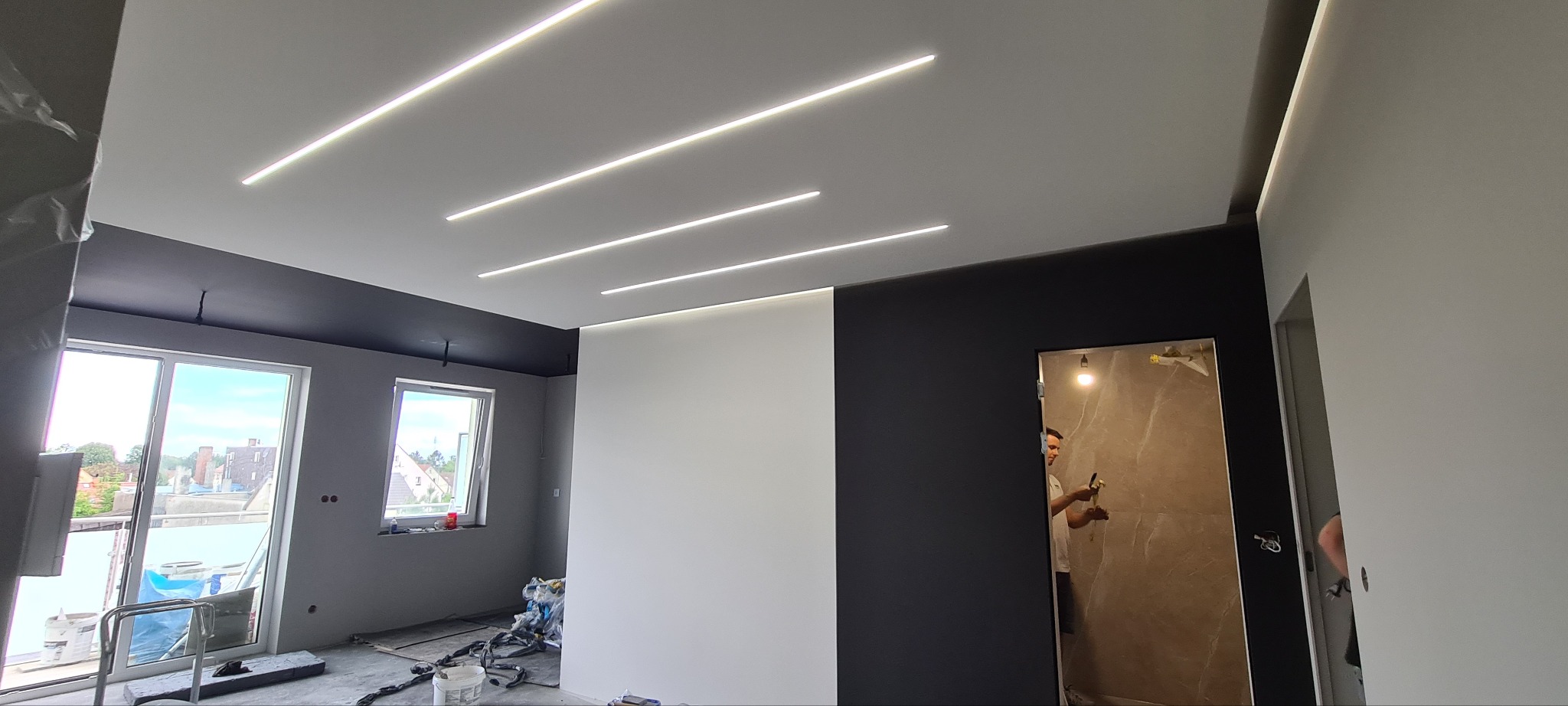 Wnętrze w trakcie wykańczania: białe ściany z ledowym oświetleniem sufitowym, czarne akcenty na suficie i ścianie, widoczne narzędzia malarskie i okna z widokiem na zewnątrz.