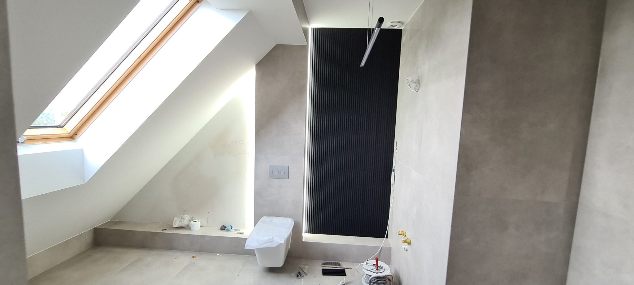 Wnętrze łazienki na poddaszu w trakcie remontu, widoczne niedokończone ściany, okno dachowe i instalacja sanitarna.