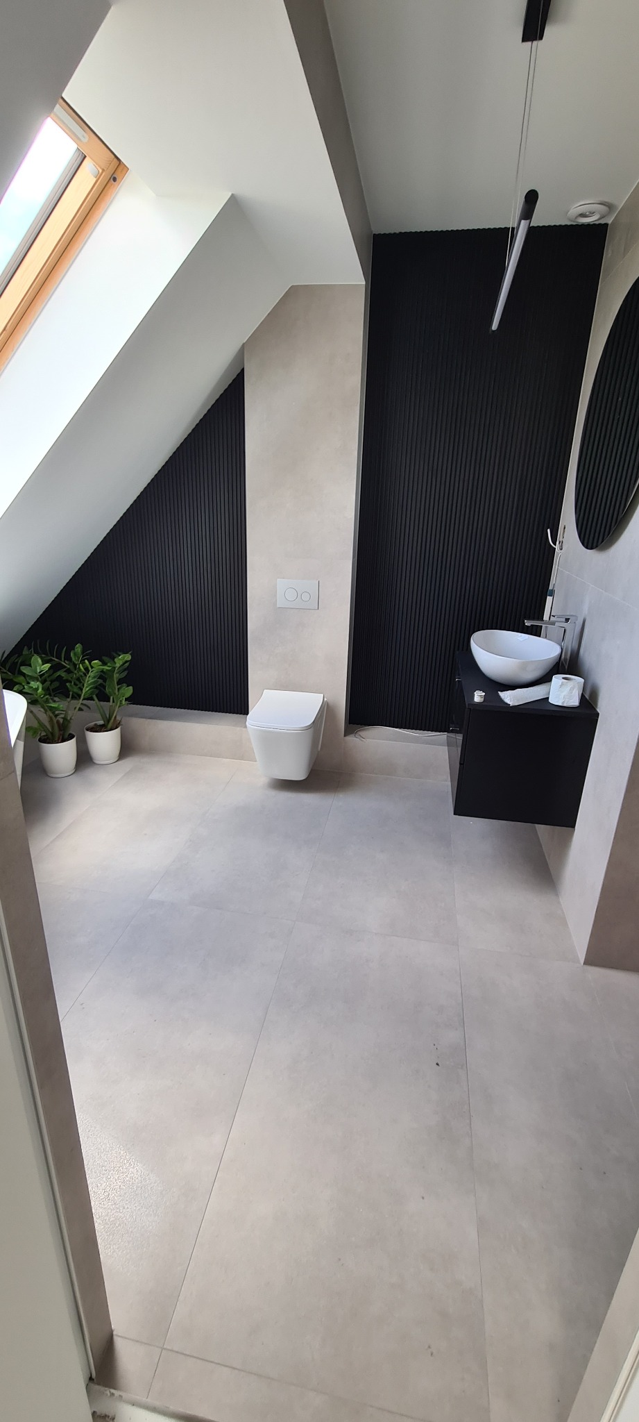 Nowoczesna łazienka na poddaszu z oknem dachowym, czarnymi panelami ściennymi, wiszącą toaletą i umywalką nablatową na czarnej szafce.