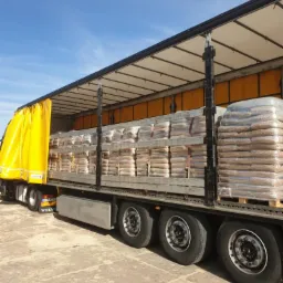 Ciężarówka z otwartą naczepą załadowana paletami z workami pelletu, zabezpieczonymi folią, widok z boku na betonowy plac.